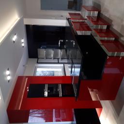 Przepis na kuchnie - Nowoczesna kuchnia z czerwonymi i czarnymi elementami, wysoka zabudowa z piekarnikiem i mikrofalówką, czarny blat i wiszące czerwone półki, oświetlenie sufitowe.