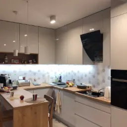 Nowoczesna kuchnia z białymi, lakierowanymi frontami szafek, drewnianym blatem i barkiem, podświetleniem LED nad blatem oraz heksagonalnymi płytkami na ścianie.