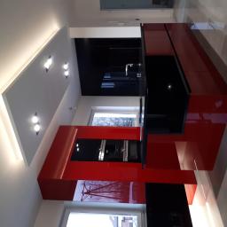 Przepis na kuchnie - Nowoczesna kuchnia z czerwono-czarnymi błyszczącymi frontami, oświetleniem sufitowym punktowym i wnękowym, widok na okno z drzewami.