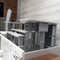 Przepis na kuchnie - Wysuwane szuflady kuchenne z organizerami na sztućce i akcesoria, widoczne w ekspozycji meblowej z jasnymi frontami i drewnianym blatem w tle.