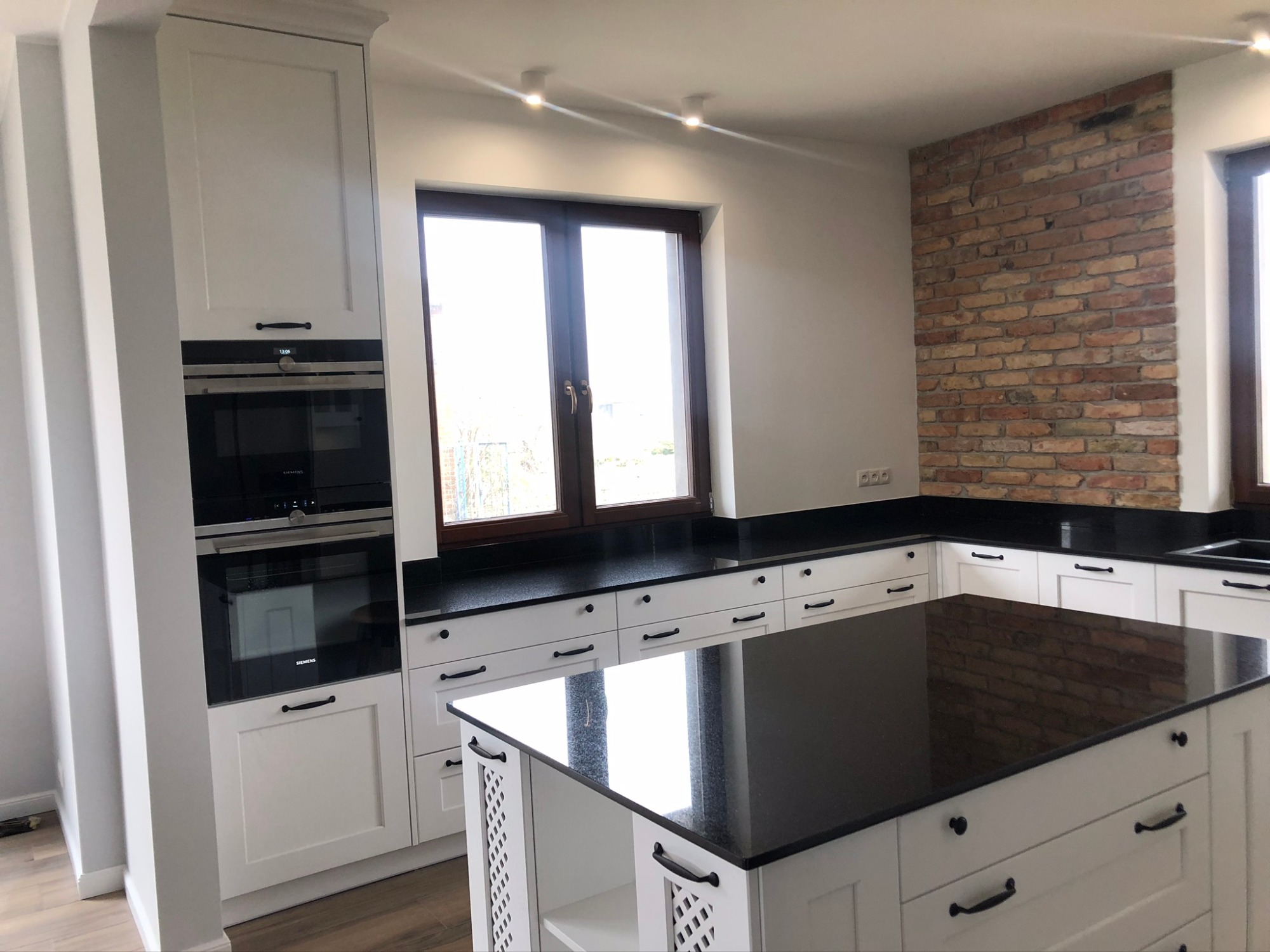 Biała kuchnia na wymiar z czarnym blatem i wyspą kuchenną, ceglana ściana w tle, nowoczesne oświetlenie punktowe.