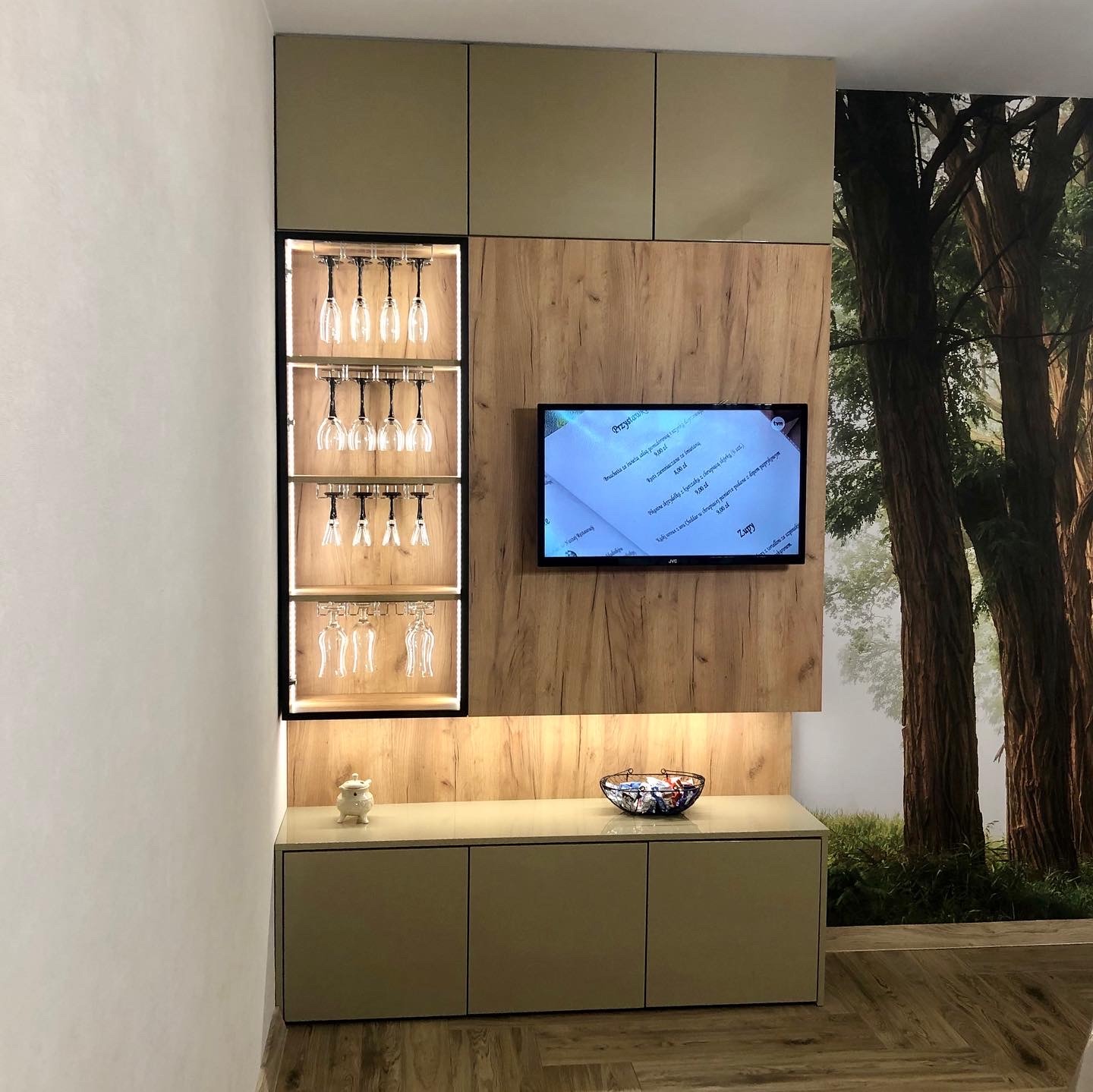 Nowoczesna zabudowa ścienna z szafkami w odcieniach beżu i drewna, z podświetlaną półką na kieliszki, telewizorem i dekoracyjną tapetą z motywem leśnym w tle.
