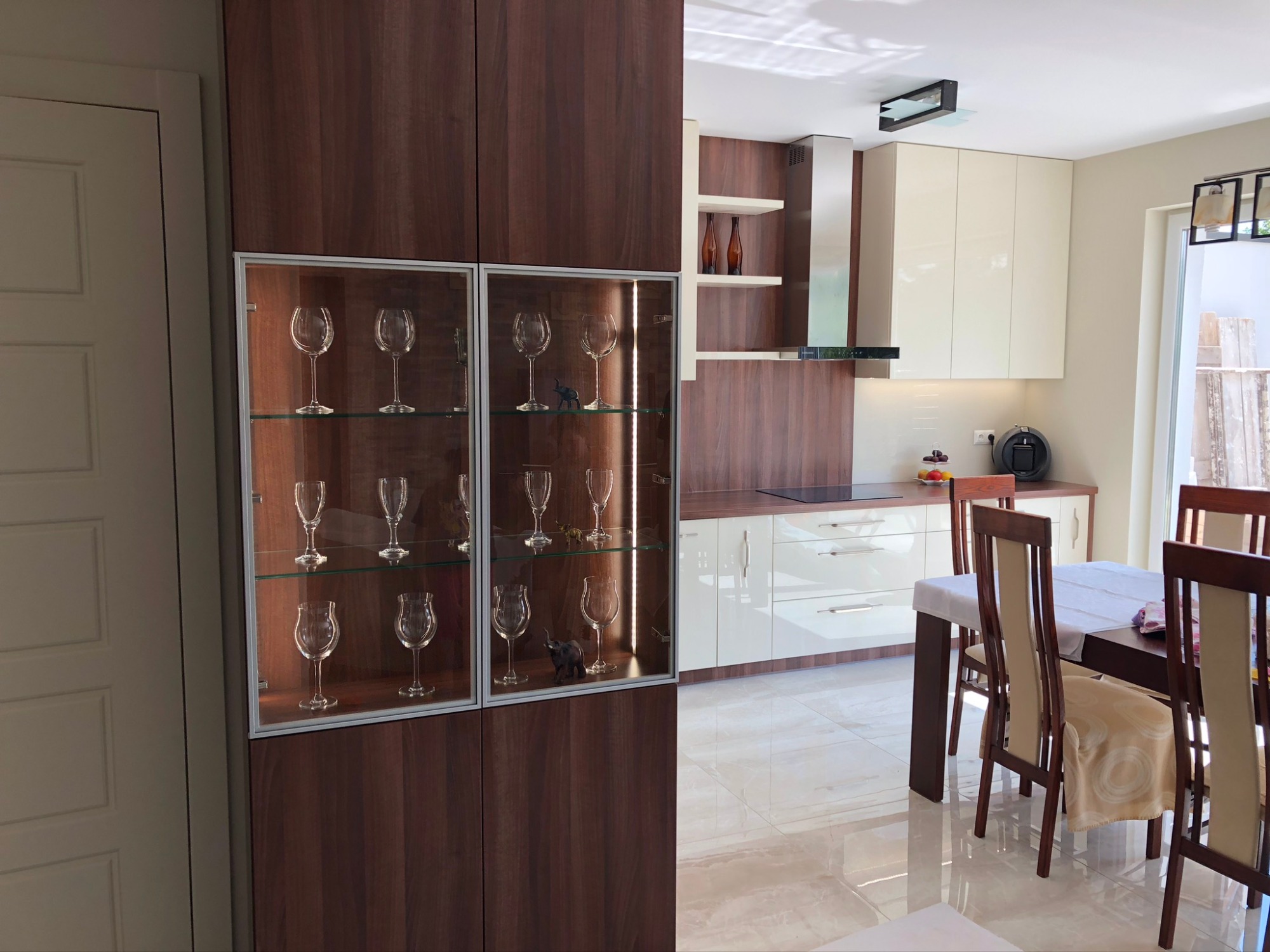 Nowoczesna kuchnia z ciemnobrązowymi szafkami, przeszkloną witryną z kieliszkami i figurką słonia, oraz jadalnią z drewnianym stołem i krzesłami w tle, oświetlona naturalnym światłem.