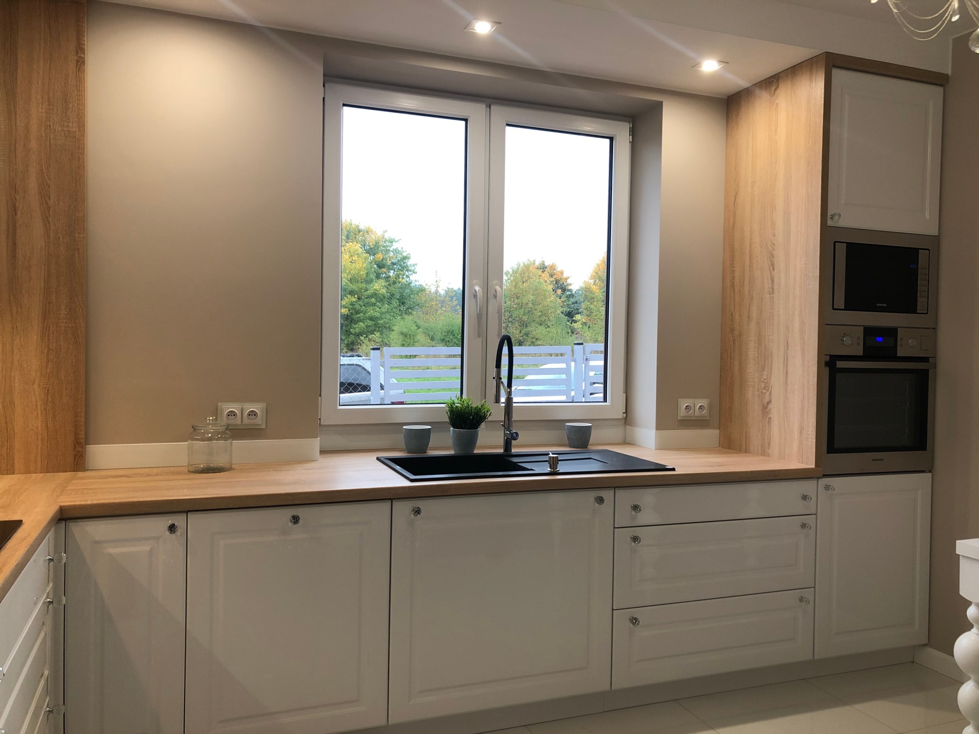 Jasna kuchnia z białymi szafkami z kryształowymi gałkami, drewnianym blatem i widokiem na zieleń za oknem.
