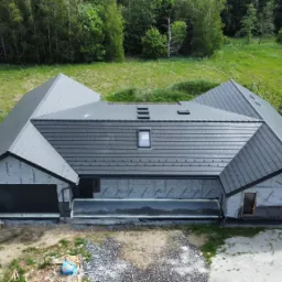 Widok z góry na dom w trakcie prac dekarskich, z szarą dachówką, oknem dachowym i folią ochronną na ścianach szczytowych, otoczony zielenią.