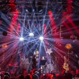 Wokalista w czerwonej cekinowej marynarce śpiewa na scenie z zespołem, oświetlonej reflektorami i kulami dyskotekowymi, widoczny tłum publiczności.