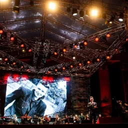 Koncert plenerowy: orkiestra symfoniczna na scenie z dużym ekranem, na którym wyświetlany jest czarno-biały portret, oświetlenie sceniczne w kolorze czerwonym i białym.