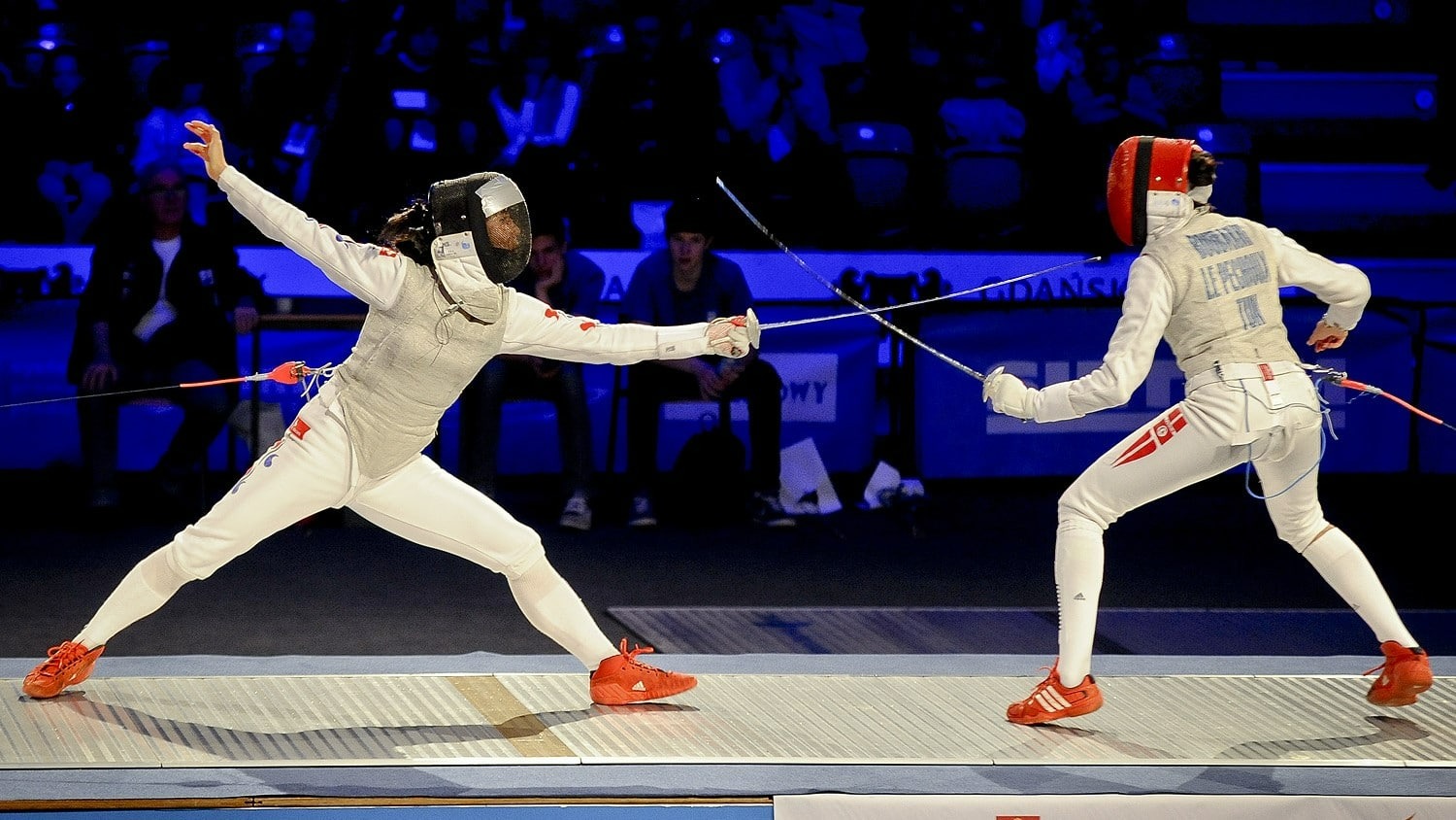 Dwóch szermierzy w białych strojach i kolorowych maskach w trakcie walki na planszy podczas zawodów sportowych, widownia w tle.