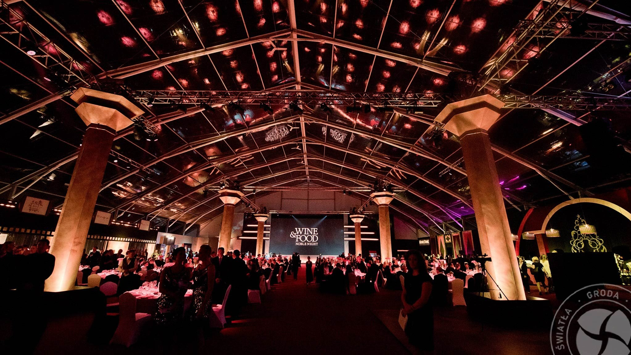 Wnętrze oświetlonego namiotu eventowego z kolumnami, przygotowanego na galę 'Wine & Food Noble Night', widoczne stoliki z gośćmi, scena z telebimem i dekoracje.