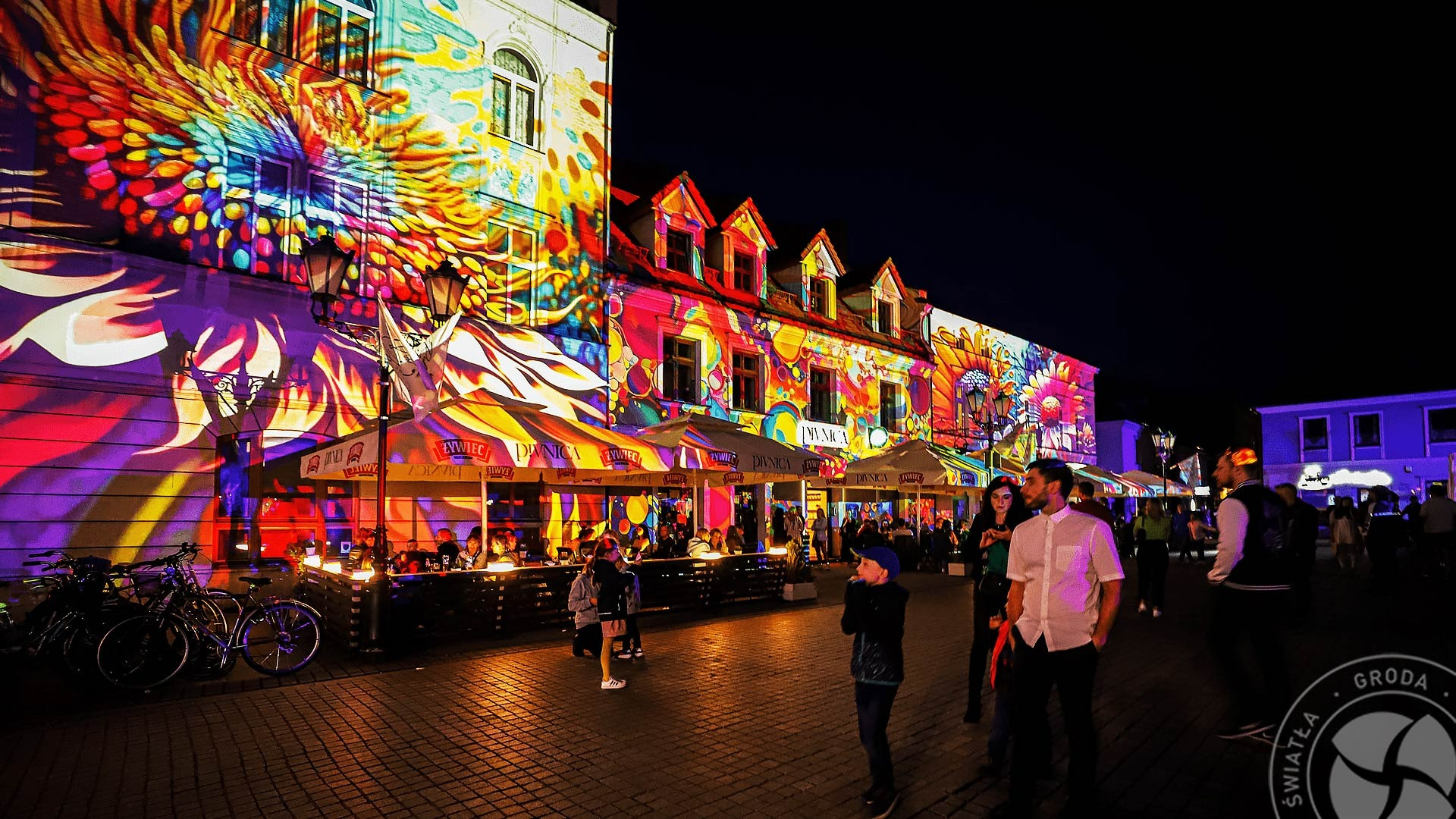Fasady budynków oświetlone kolorowymi projekcjami świetlnymi podczas nocnego wydarzenia plenerowego, widoczne stoliki kawiarniane pod parasolami i spacerujący ludzie.