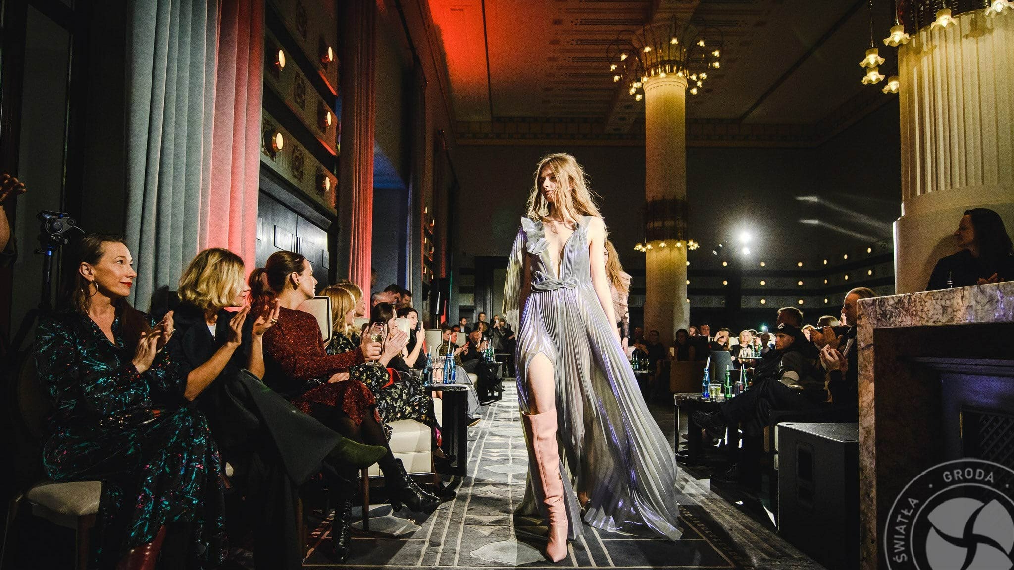 Modelka w srebrnej sukni z rozcięciem prezentuje się na wybiegu podczas pokazu mody, publiczność obserwuje i klaszcze, oświetlenie sceniczne w odcieniach czerwieni i bieli, luksusowe wnętrze...