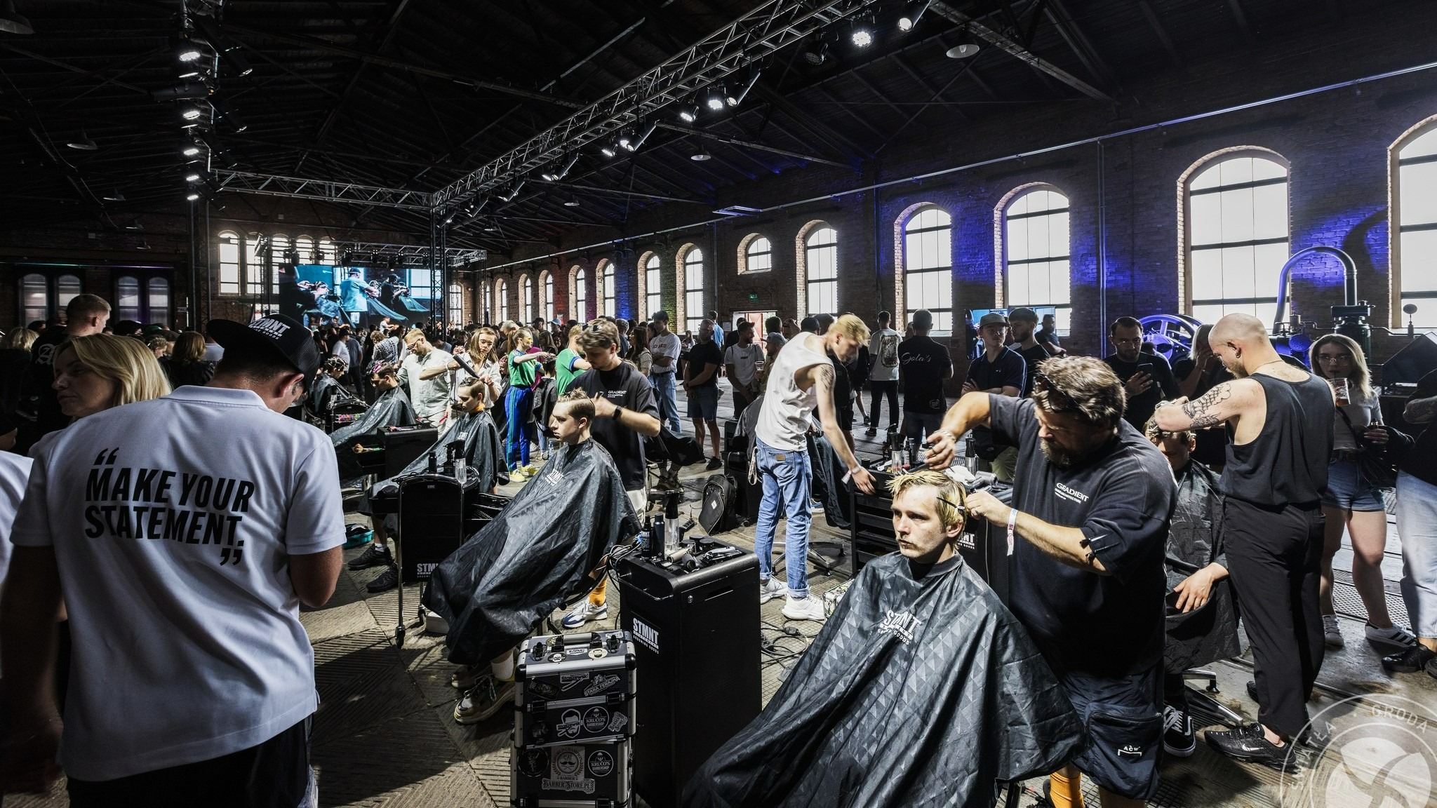 Salon fryzjerski na evencie, fryzjerzy strzygą klientów w dużym, industrialnym pomieszczeniu z ceglanymi ścianami i oknami łukowymi, oświetlonym reflektorami.