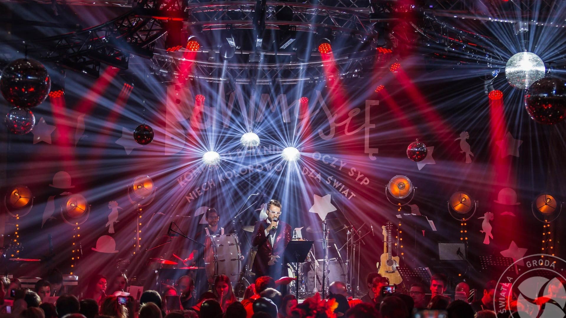 Wokalista w czerwonej cekinowej marynarce śpiewa na scenie z zespołem, oświetlonej reflektorami i kulami dyskotekowymi, widoczny tłum publiczności.
