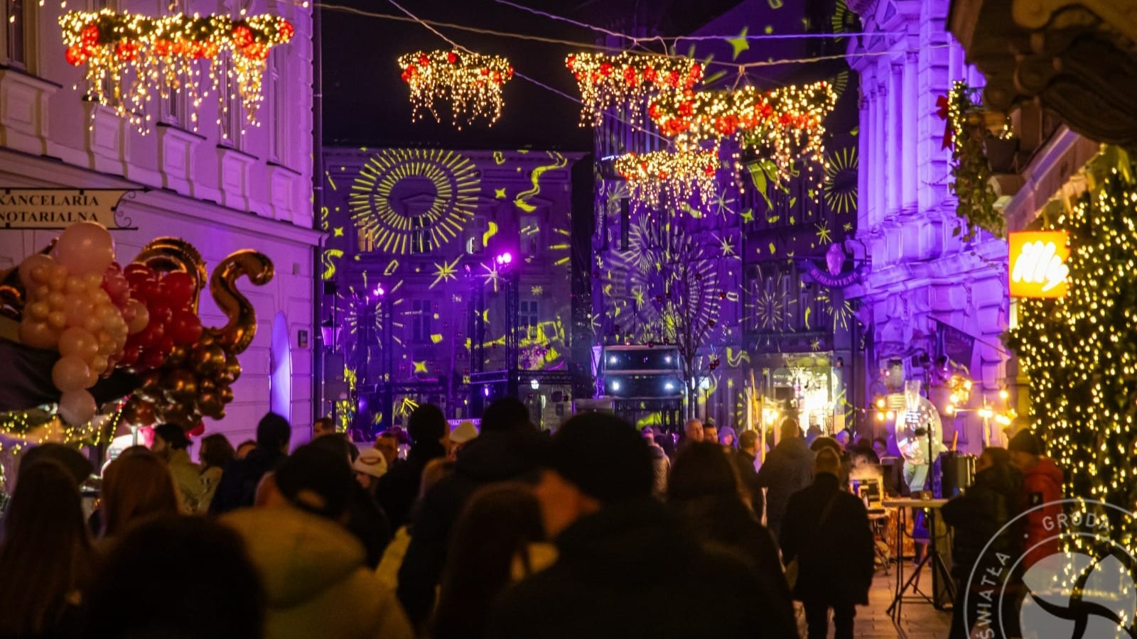 Ulica nocą z dekoracjami świątecznymi, tłum ludzi, budynki oświetlone projekcjami świetlnymi w kolorze fioletowym i żółtym, girlandy z czerwonymi ozdobami i białymi światełkami, balony dekoracyjne...