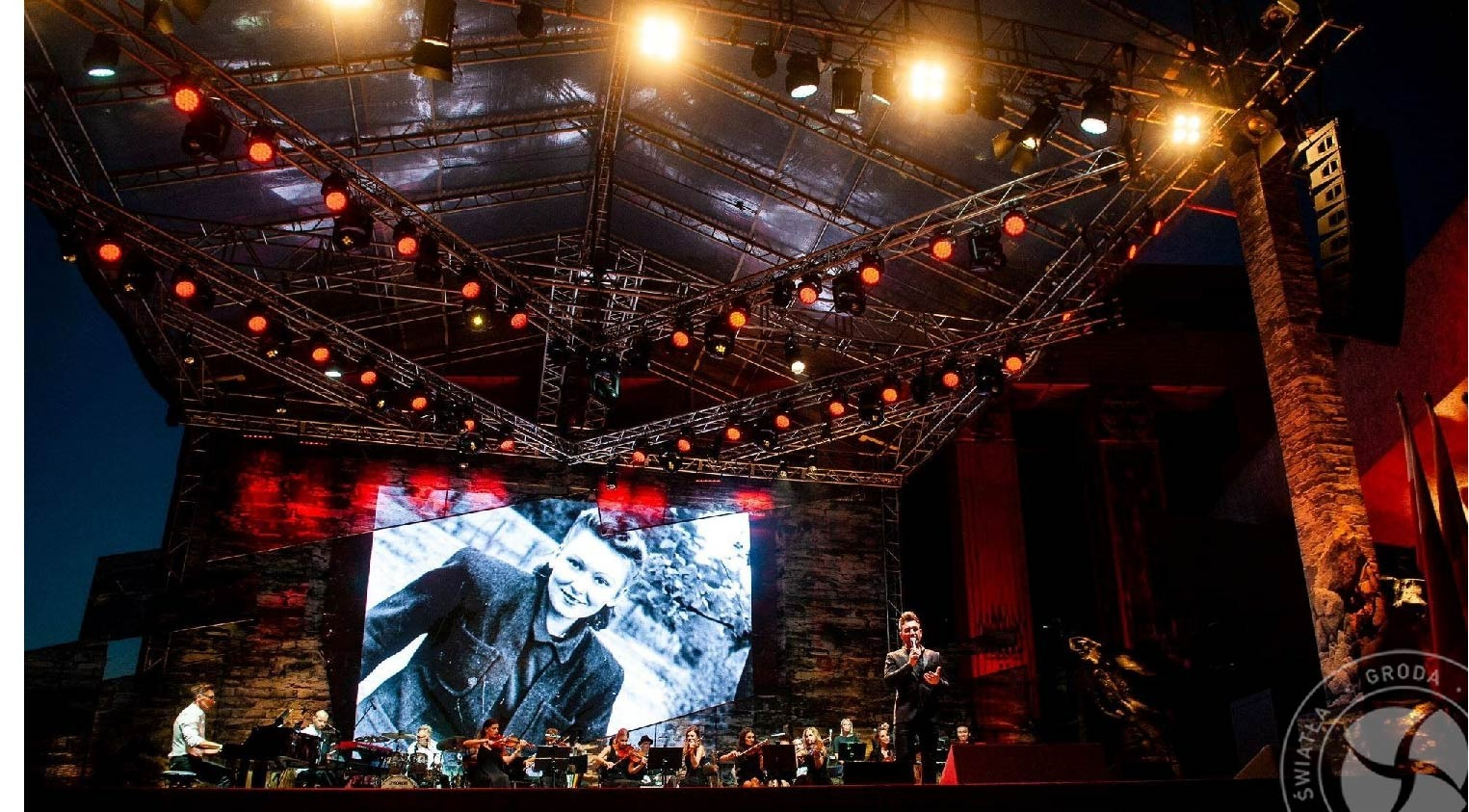 Koncert plenerowy: orkiestra symfoniczna na scenie z dużym ekranem, na którym wyświetlany jest czarno-biały portret, oświetlenie sceniczne w kolorze czerwonym i białym.
