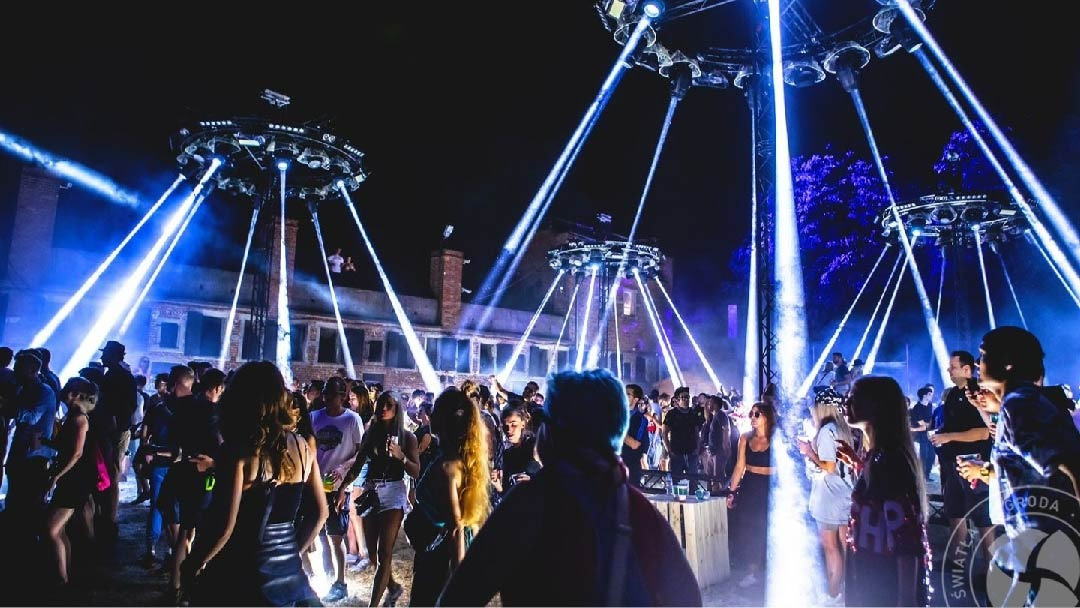 Tłum ludzi bawi się na imprezie plenerowej oświetlonej przez trzy okrągłe konstrukcje z reflektorami emitującymi intensywne niebieskie światło, w tle budynek z cegły.