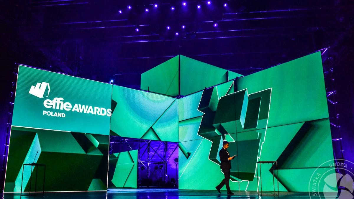 Scena z zielono-niebieskim oświetleniem podczas gali Effie Awards Poland, z prelegentem przechodzącym obok podświetlanych ekranów.