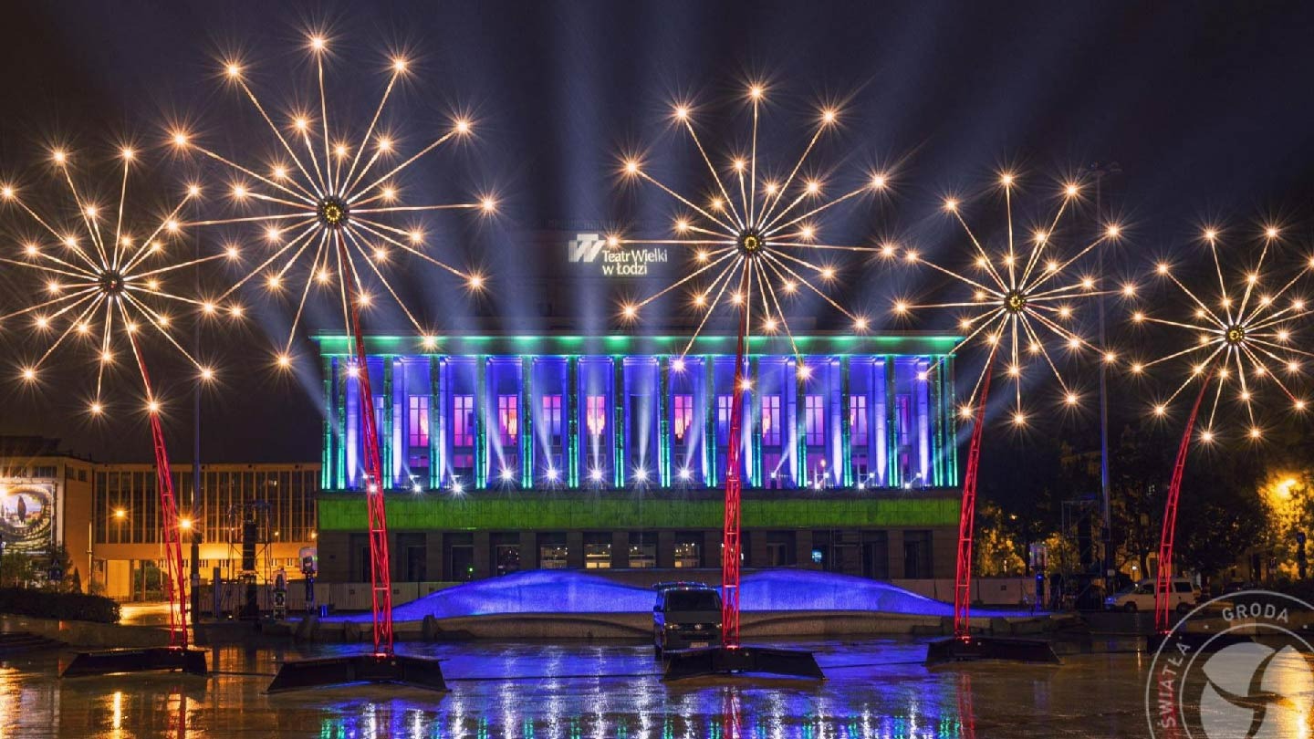 Teatr Wielki w Łodzi nocą, oświetlony dekoracyjnymi świetlnymi konstrukcjami w kształcie gwiazd, odbijającymi się w wodzie.