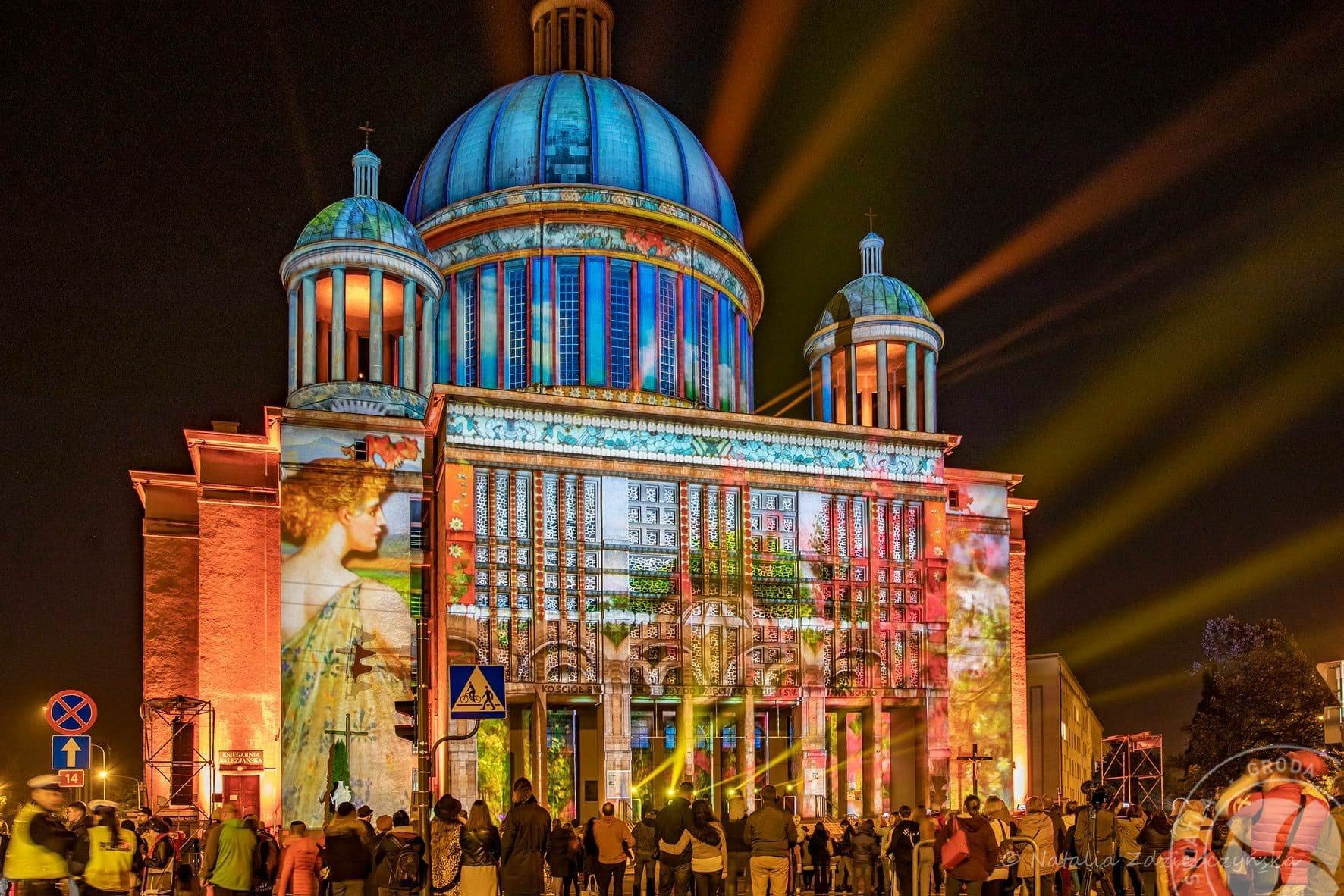 Iluminacja budynku z kopułą, wyświetlane obrazy i wzory, tłum obserwatorów w nocy, promienie światła przecinające ciemne niebo.