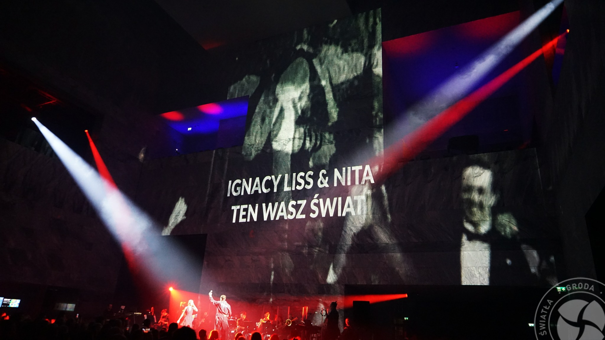 Koncert z orkiestrą na scenie, z projekcją w tle z napisem 'Ignacy Liss & Nita Ten Wasz Świat', oświetlony reflektorami w kolorze białym i czerwonym.
