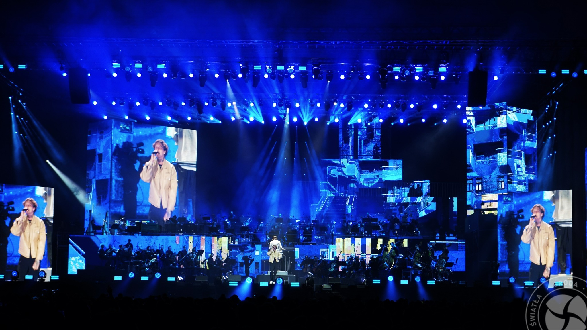 Scena koncertowa z wokalistą i orkiestrą symfoniczną, oświetlona intensywnym niebieskim światłem, z ekranami LED wyświetlającymi obraz wokalisty i abstrakcyjne wizualizacje.