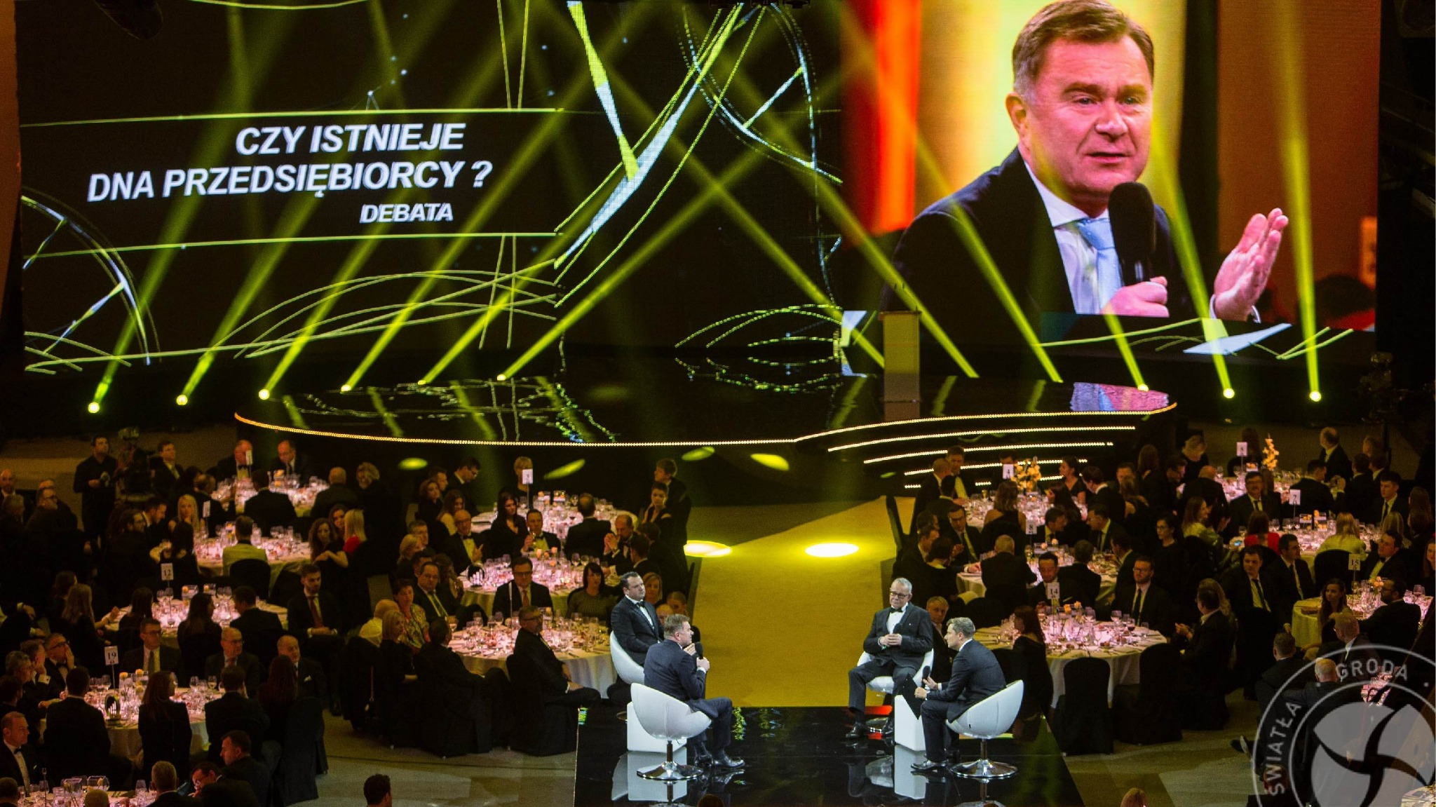 Sala konferencyjna wypełniona gośćmi, na scenie debata z oświetleniem punktowym i projekcją wizerunku prelegenta na telebimie, stoliki z numerami dla uczestników.