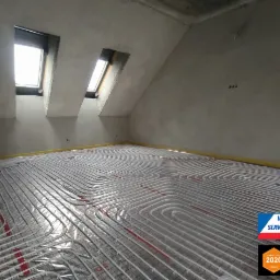 Białe rury instalacji grzewczej podłogowej ułożone na folii z czerwoną siatką, widoczne dwa okna dachowe w surowym pomieszczeniu ze ścianami w szarym tynku, w rogu naklejka z logo firmy i nagrodą...