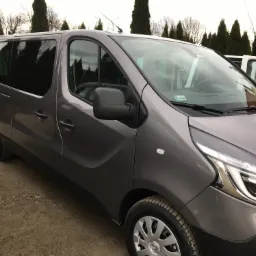 Szary, nowoczesny van pasażerski z przyciemnianymi szybami, zaparkowany na szutrowym podłożu, z widoczną anteną na dachu i zawieszką zapachową wewnątrz.