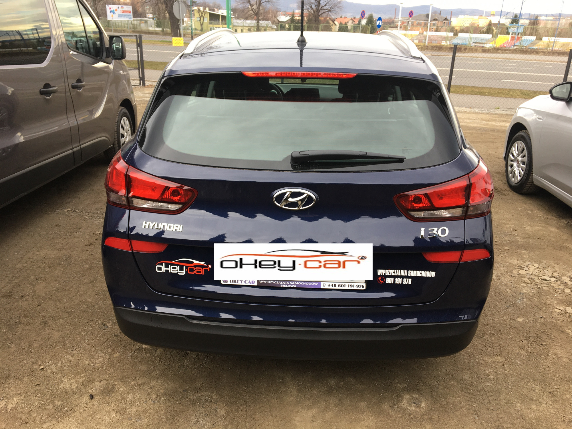 Tył granatowego samochodu Hyundai i30 kombi z naklejkami firmy 'okey-car' oferującej wynajem aut, zaparkowanego na żwirowym parkingu.