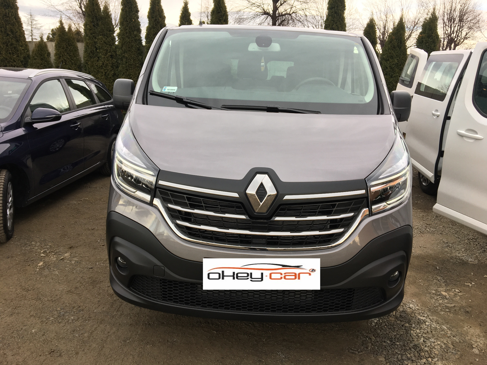 Szary Renault Trafic z przodu, z widocznym logo firmy okey-car.pl na dolnej części zderzaka, stojący na żwirowym podłożu obok innych pojazdów.