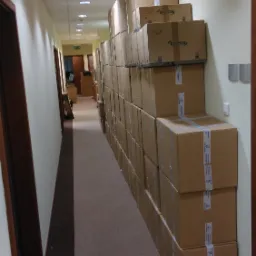 Długi korytarz biurowy zastawiony wysokim rzędem kartonowych pudeł, częściowo zaklejonych taśmą, niektóre z nadrukowanym logo Lyreco, oświetlony punktowymi lampami sufitowymi.