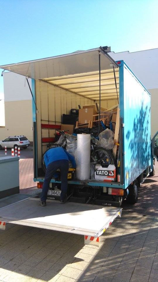 Otwarta paka dostawczego samochodu wypełniona kartonami, meblami i rowerami, zabezpieczonymi folią, z widocznym pracownikiem w niebieskiej bluzie.