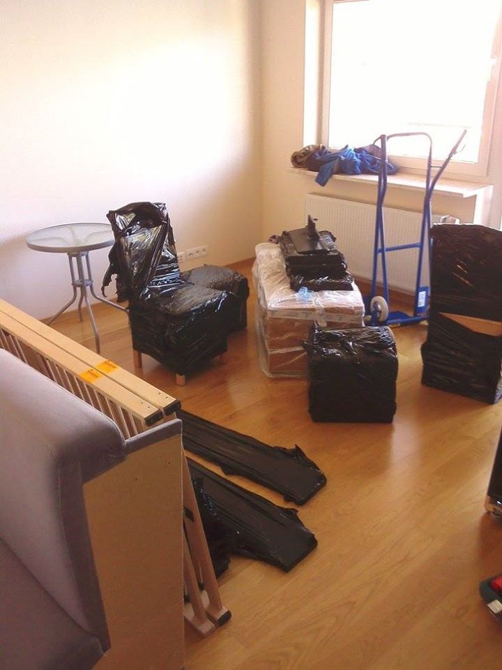 Pokój z przygotowanymi do transportu meblami, zabezpieczonymi czarną folią, w tym krzesło, stolik i elementy sofy, oraz wózkiem transportowym w tle przy oknie.