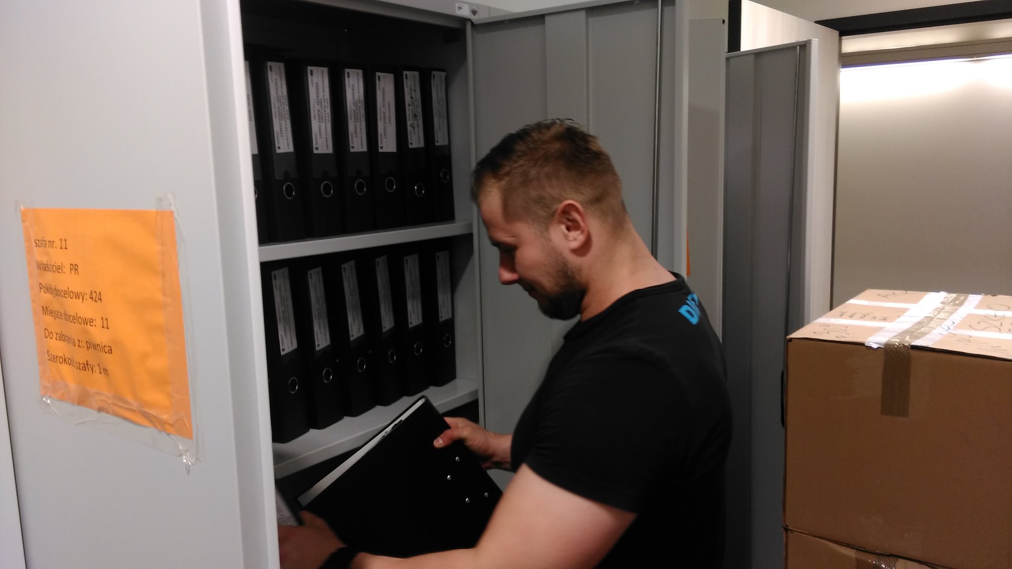Mężczyzna wkłada czarny segregator do metalowej szafy z rzędami segregatorów, na drzwiach szafy żółta kartka z napisem 'szafa nr. 11, do zabrania z piwnicy', obok kartonowe pudła.
