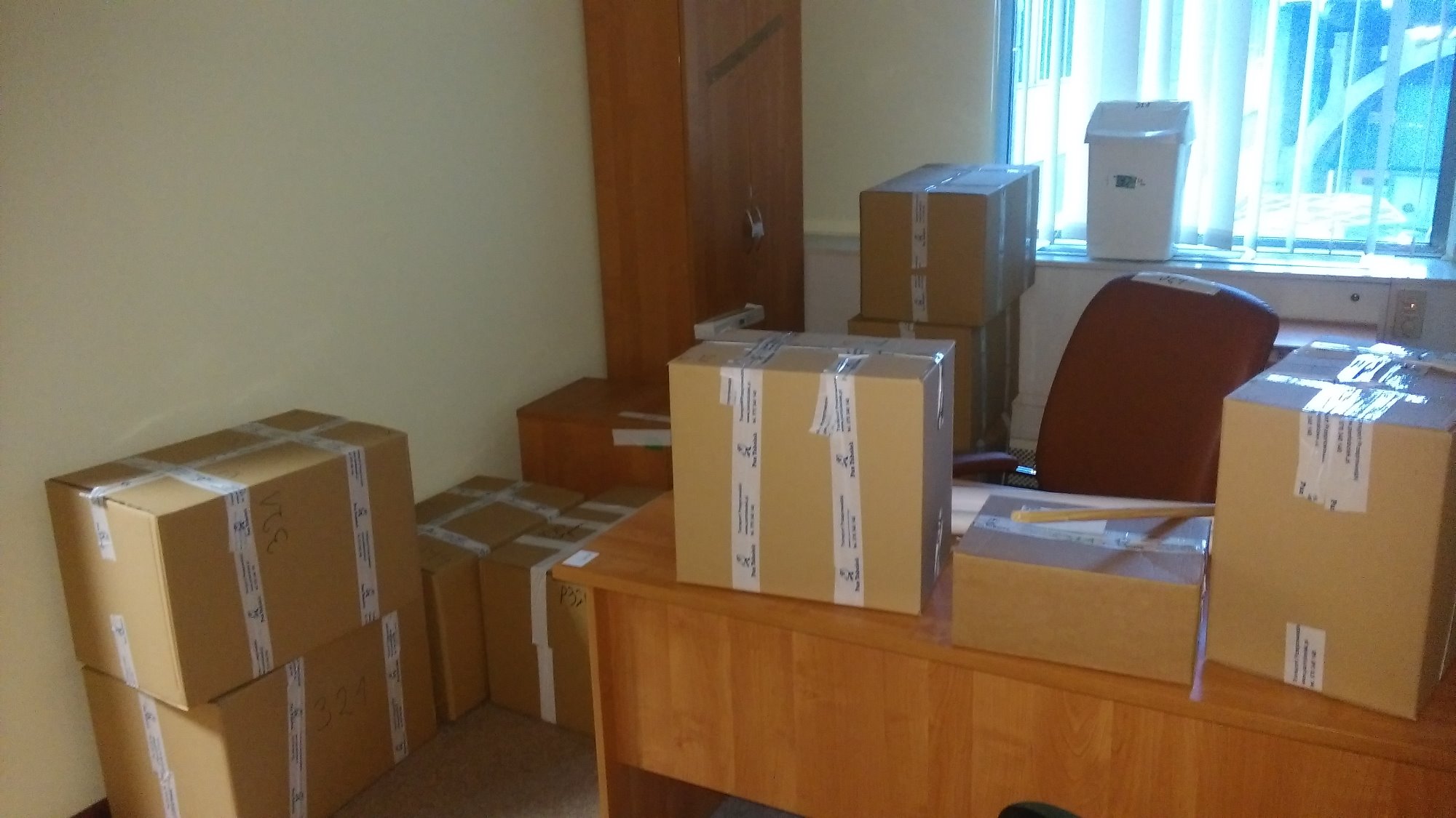 Kartonowe pudła z taśmą zabezpieczającą, niektóre z numerami '321', ustawione w biurze przed oknem z żaluzjami, na biurku i podłodze, przygotowane do transportu.