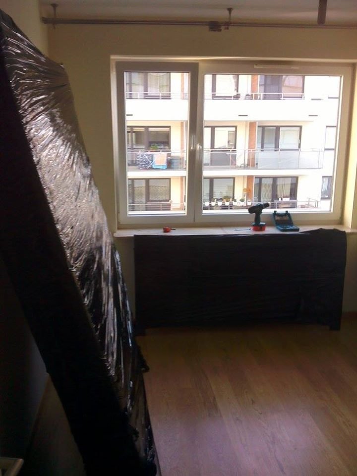 Pionowo ustawiony, owinięty w folię mebel w pustym pokoju z widokiem na budynek mieszkalny; w tle wiertarka i akcesoria na parapecie.