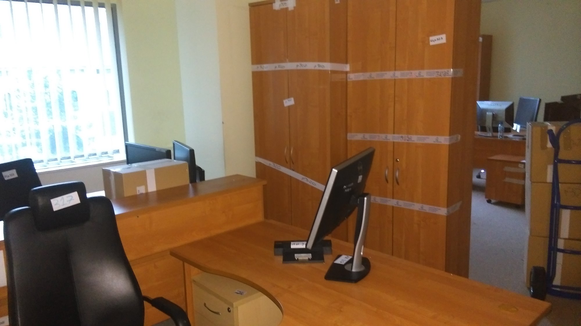 Biuro przygotowane do transportu: spakowane szafy oklejone taśmą, biurka z monitorami, krzesła z numerami, kartonowe pudła i wózek transportowy.