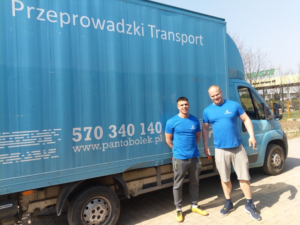 Dwóch uśmiechniętych mężczyzn w niebieskich koszulkach stoi przed niebieską ciężarówką z napisem 'Przeprowadzki Transport' i numerem telefonu, zaparkowaną na chodniku.