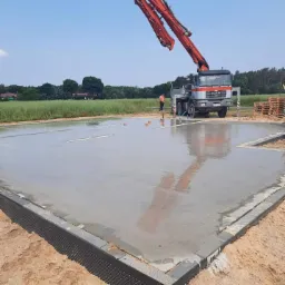 Wylewanie betonu na przygotowany fundament z izolacją pionową, widoczny samochód z pompą do betonu i pracownik w oddali.