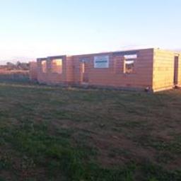 Usługi Budowlane TOMKOWICZ Weronika Tomkowicz - Wczesny etap budowy domu z bloczków, widoczne ściany bez dachu i okien, na ścianie widnieje tablica informacyjna budowy, w tle pole i niebo.