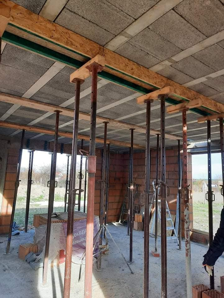 Wnętrze w trakcie budowy: widoczne ceglane ściany, betonowy strop podparty metalowymi stemplami i drewnianymi belkami, z zapasem materiałów budowlanych w tle.