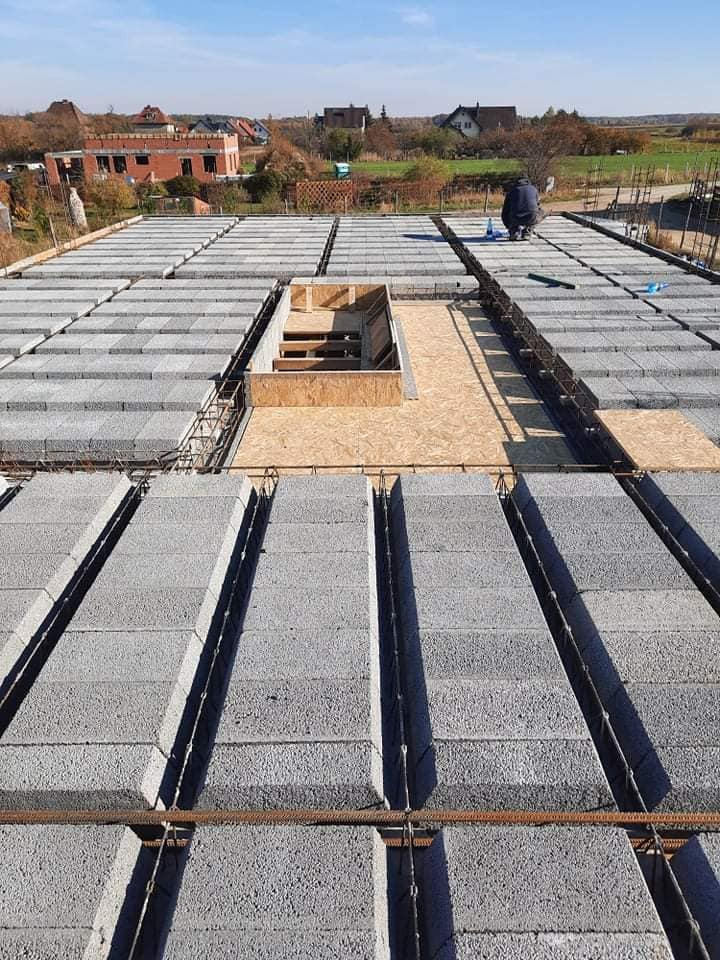 Wznoszenie stropu z pustaków szalunkowych; widoczny otwór na schody zabezpieczony drewnianym szalunkiem i pracownik w oddali.