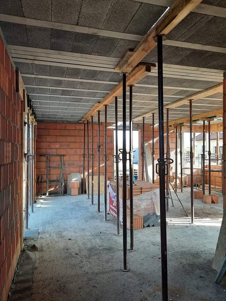 Wnętrze budynku w trakcie budowy z widocznymi ścianami z cegły, betonowym stropem podpartym metalowymi stemplami oraz materiałami budowlanymi na podłodze.