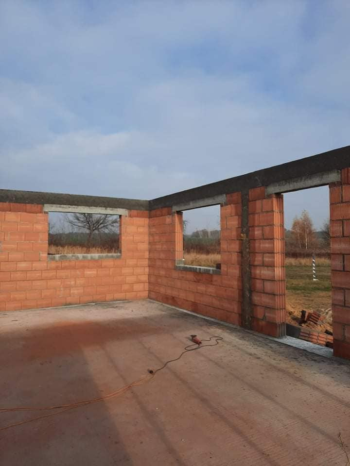 Wnętrze surowego domu murowanego z czerwonej cegły z widocznymi otworami okiennymi i betonowym nadprożem, na podłodze leży przewód elektryczny