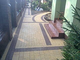 Ułożona ścieżka z kostki brukowej w dwóch odcieniach, z ciemniejszym obramowaniem i łukowatym wzorem, prowadząca wzdłuż ceglanego muru z drewnianymi siedziskami do wejścia budynku z brązowymi...