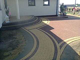 Ułożona kostka brukowa w kształcie łuków prowadząca do domu, widoczna maszyna do zagęszczania i pracownik.