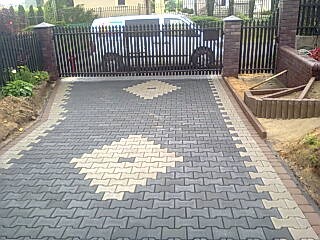 Ułożona kostka brukowa w geometryczny wzór na podjeździe, z widoczną bramą w tle i przygotowanym obrzeżem po prawej stronie.