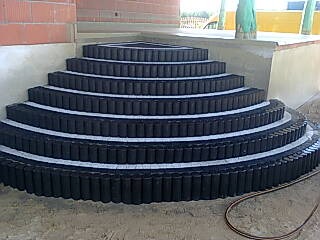 Półokrągłe schody zewnętrzne wykonane z czarnej kostki o cylindrycznym kształcie z białymi fugami, prowadzące do wejścia budynku.