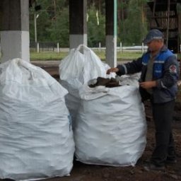 PHU BIALENERGO Olga Choińska - Mężczyzna w uniformie sprawdza zawartość worka z ciemnym materiałem na tle trzech dużych, białych worków pod zadaszeniem.