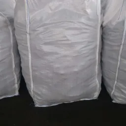 Duże, białe worki z zawartością, ułożone obok siebie, z widocznymi uchwytami do przenoszenia i niebieskimi szwami, stojące na ciemnej, prawdopodobnie betonowej powierzchni.